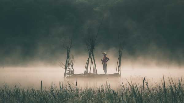 Pria dengan topi berdiri di atas perahu di Danau Tamblingan, dikelilingi oleh kabut pagi dan latar belakang hutan.