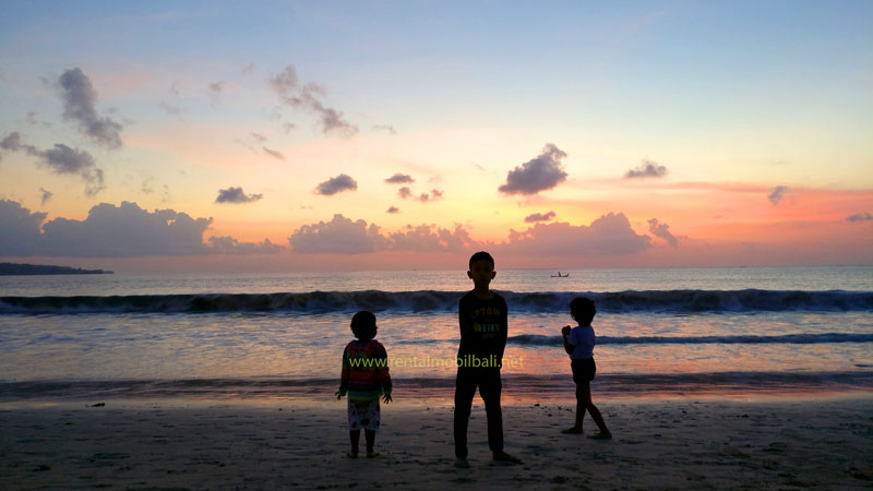 Tiga anak riang bermain di pantai pasir putih Jimbaran saat matahari terbenam yang cerah di Bali.