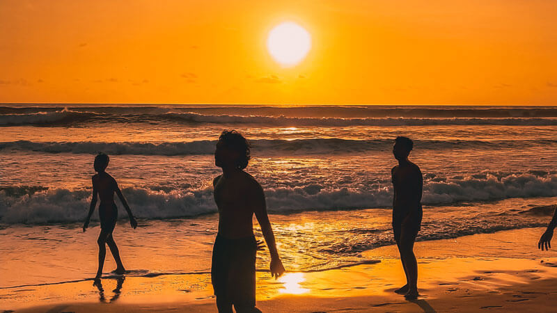 Matahari terbenam yang mempesona di Pantai Seminyak dengan siluet orang-orang yang menikmati ombak. Panduan Wisata Lengkap ke Seminyak