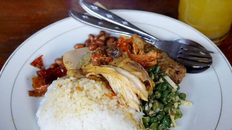 Piring nasi putih dengan lauk ayam betutu, lawar, dan kacang, serta sambal di samping minuman kuning