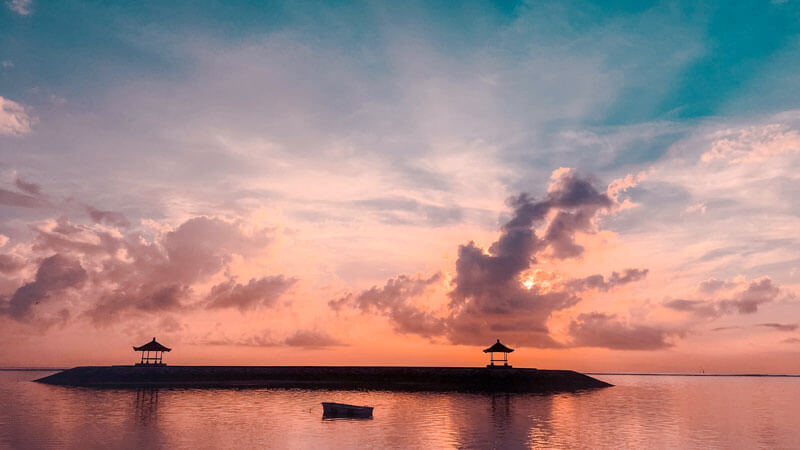 Sunrise view di Bali dengan langit berwarna jingga, pantulan cahaya matahari di permukaan air yang tenang, dan dua gazebo di ujung tanggul. Pertanyaan Umum Liburan di Bali