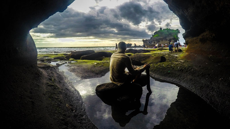 Seorang pengunjung merenung sambil duduk di batu, menghadap Pura Tanah Lot di sore hari dengan awan mendung di atasnya.