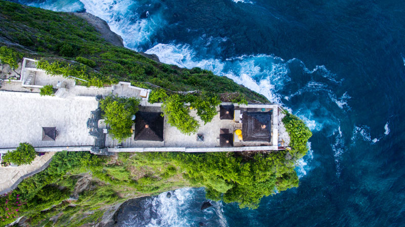 Pemandangan udara Pura Uluwatu di atas tebing dengan latar belakang Samudra Hindia di Bali, menggambarkan kedekatan pura dengan alam dan suasana spiritualnya.