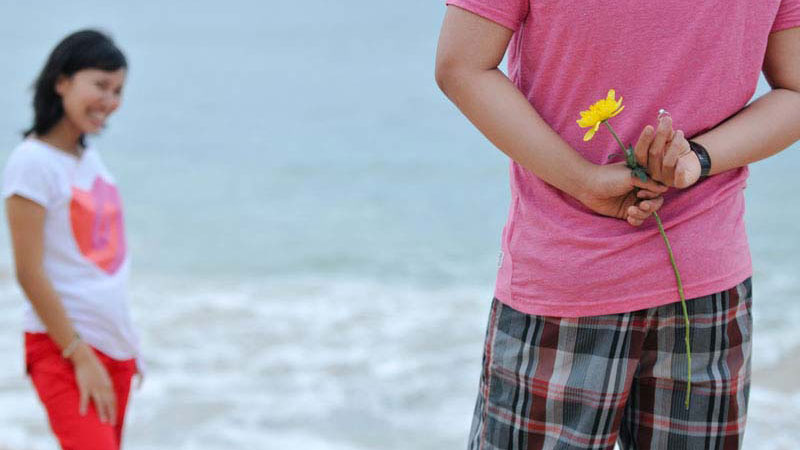 Pria dengan kemeja pink menyembunyikan bunga di belakang punggung di pantai, dengan wanita tersenyum di latar belakang