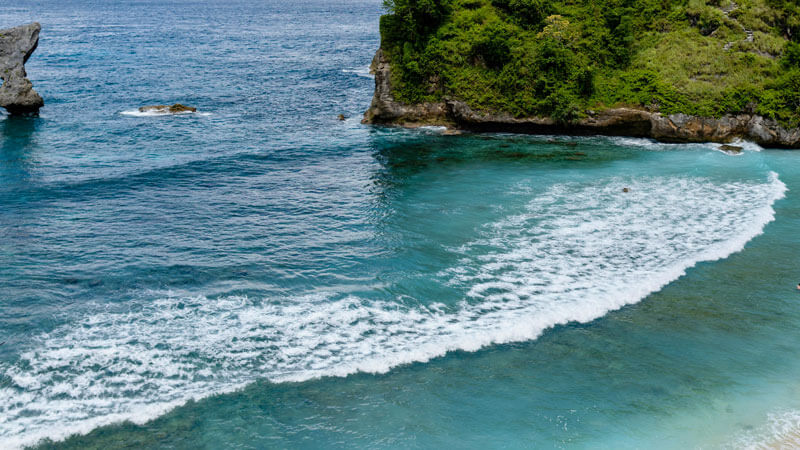 Pemandangan Pantai Atuh di Nusa Penida dengan ombak berbusa di tepi pasir putih dan tebing hijau di sekelilingnya