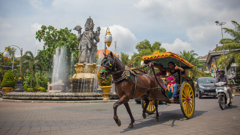 Delman tradisional melintas di depan Monumen Bajra Sandhi dengan patung heroik di latar belakang dan semburan air mancur, Denpasar, Bali