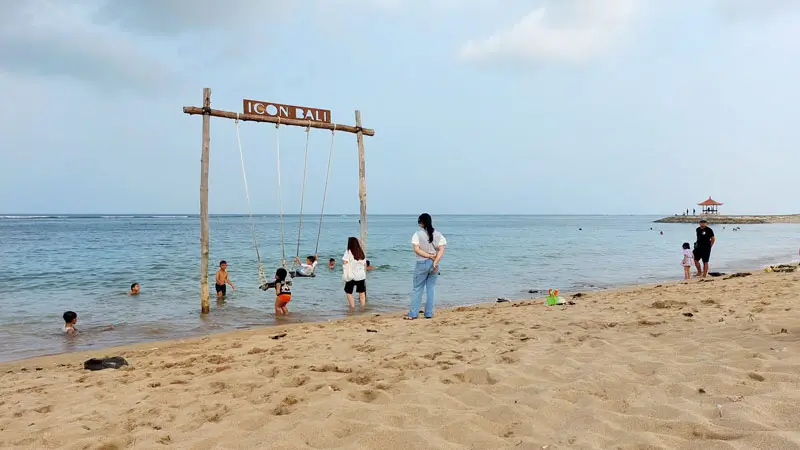 Anak-anak bermain ayunan di pantai Sanur dengan suasana santai dan ramah keluarga.