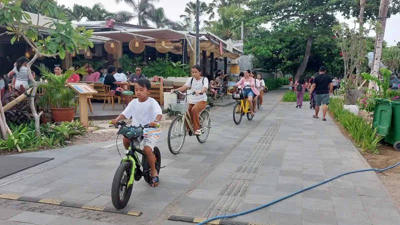 Keluarga bersepeda di promenade Tempat Wisata Sanur dengan latar restoran dan jalur hijau.