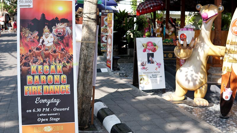 Banner promosi Pertunjukan Tari Kecak dan Barong dengan latar senja dan patung karakter komik di Tanah Lot Bali.