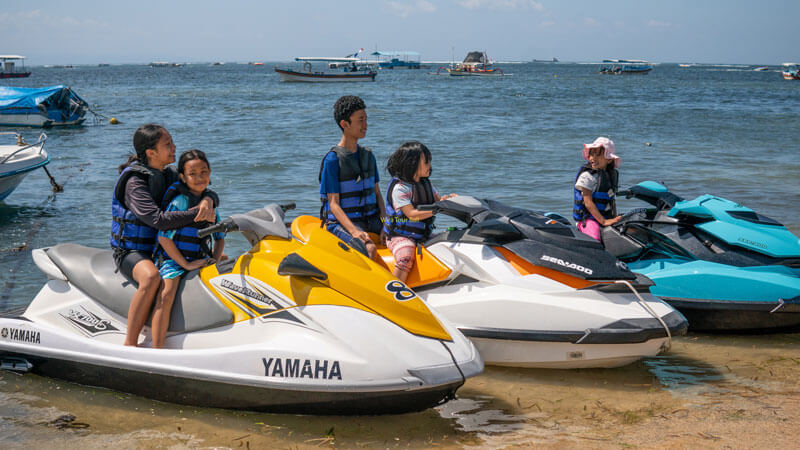 Keluarga bersiap untuk menaiki jet ski di pantai Bali dengan pemandangan kapal-kapal di latar belakang