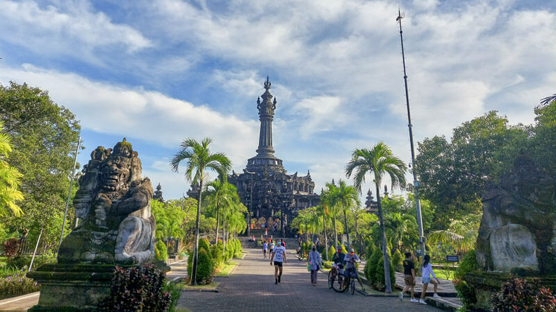 Pemandangan Monumen Bajra Sandhi di Bali pada siang hari dengan langit biru dan pengunjung yang berjalan di jalur taman.