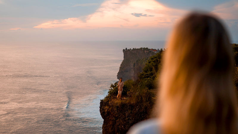 Seorang pengunjung mengagumi pemandangan matahari terbenam dari tebing Pura Uluwatu, Bali, dengan samudra yang luas di latar belakang.