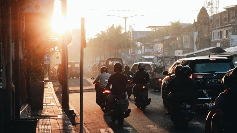 Kendaraan bermotor dan mobil melaju di jalan Bali saat matahari terbenam, menggambarkan perjalanan menuju Pura Luhur Uluwatu.