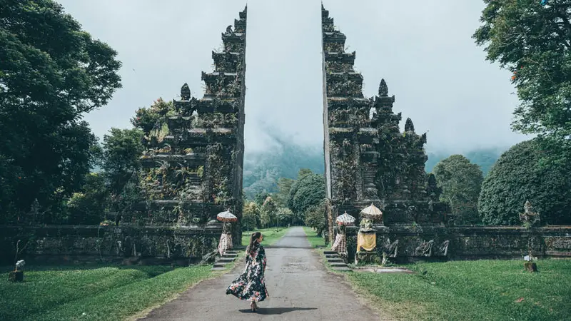 Wisatawan berjalan mengagumi gerbang candi kuno di Bali, berlatar kabut pegunungan.