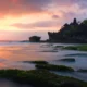 Pura Tanah Lot Bali saat matahari terbenam, pemandangan indah dengan ombak dan batu karang di sekitarnya.