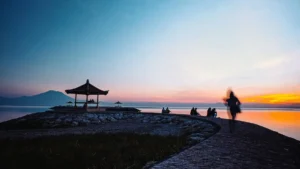 Wisata sunrise di Pantai Sanur Denpasar dengan gazebo dan beberapa pengunjung di tepi laut tenang